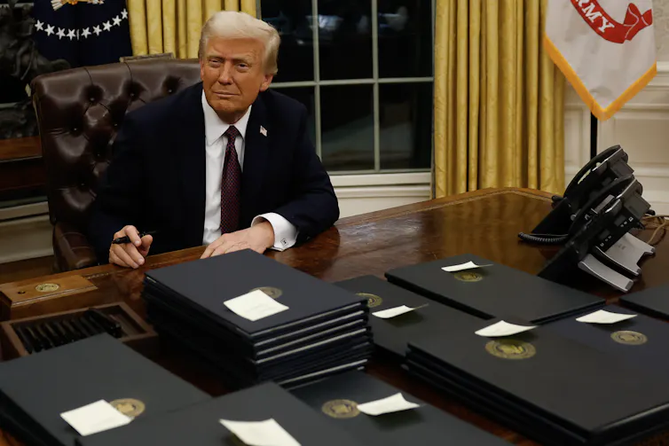 A light-haired man in a suit holding a pen at a desk covered with folders.