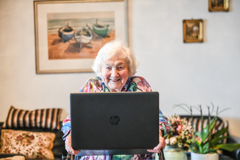 An older woman with white hair smiles and looks over the screen of a laptop.