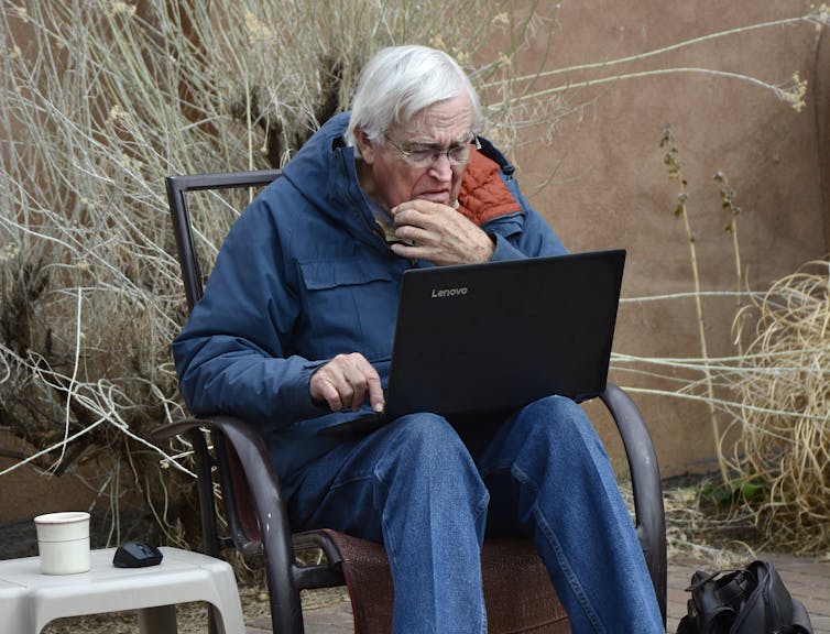An older man with white hair sits outside and looks at the screen of a laptop.