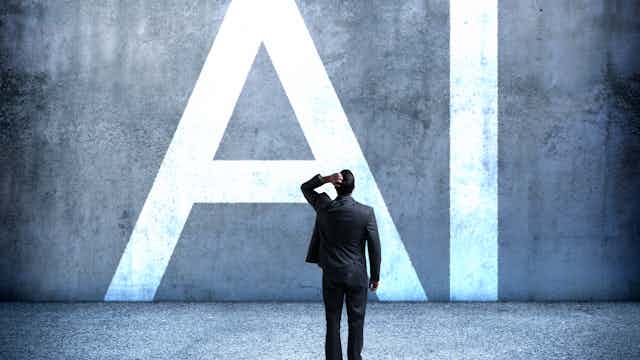 View from the back of a man wearing a business suit with his left hand on the back of his head facing a concrete wall with giant letters 'AI' on it