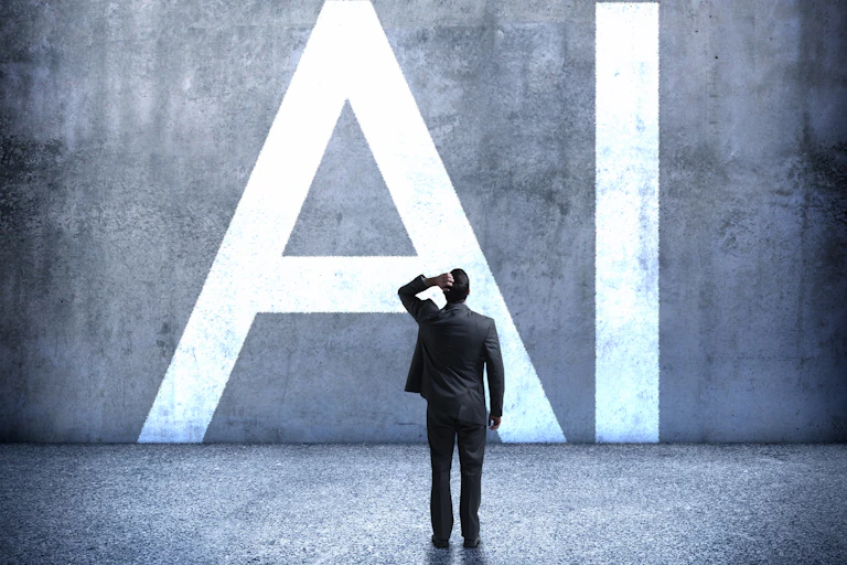 View from the back of a man wearing a business suit with his left hand on the back of his head facing a concrete wall with giant letters 'AI' on it