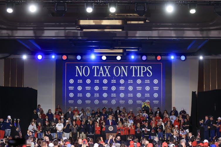 Donald Trump stands in front of a very large blue sign that reads 'No tax on tips.'