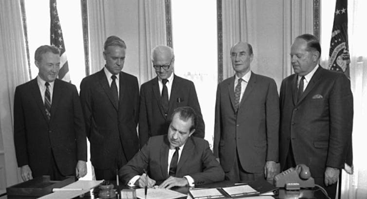 Man seated at desk writing on papers with five men in suits behind.