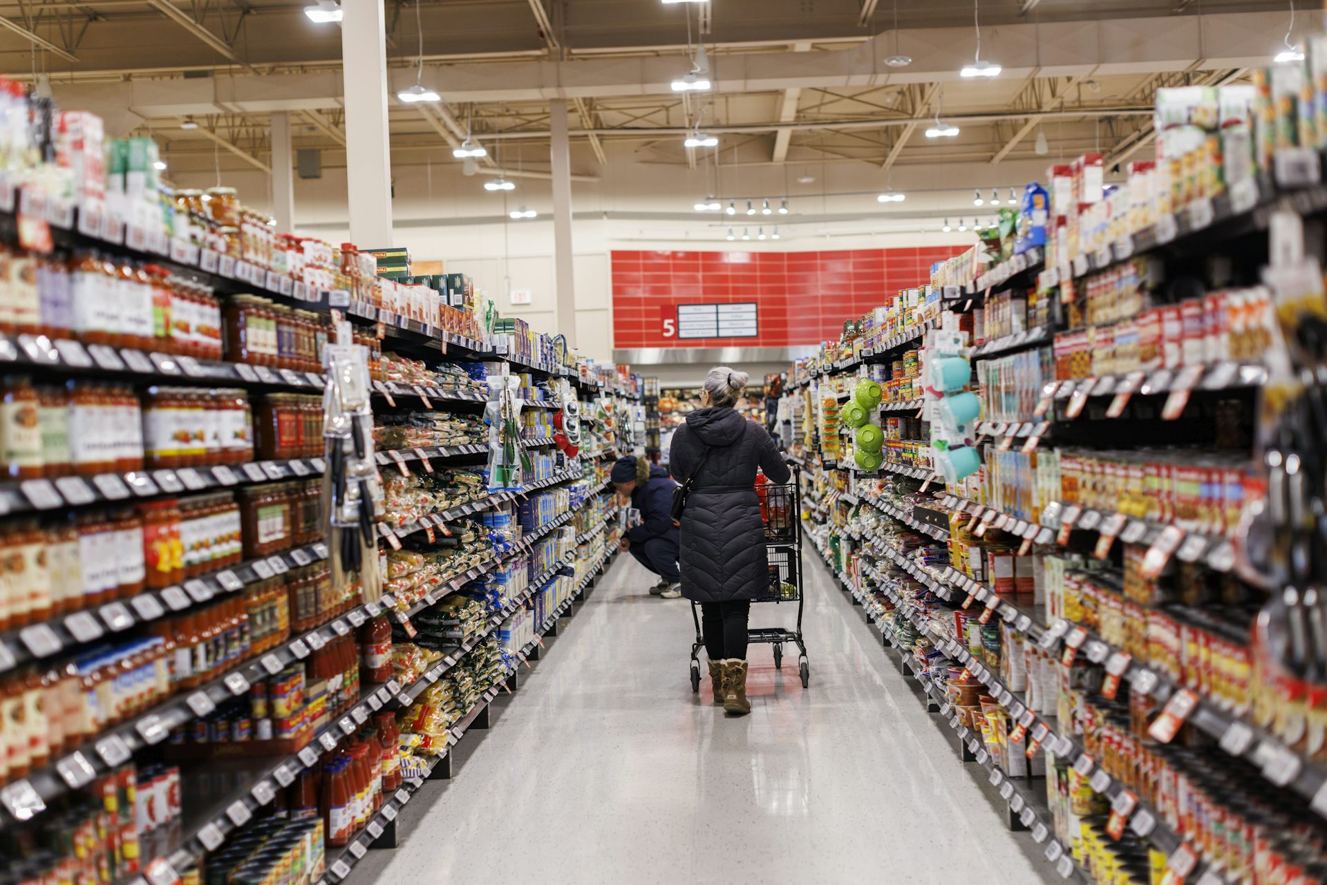 A woman seen from behind pushing a grocery cart down an aisle in a supermarket