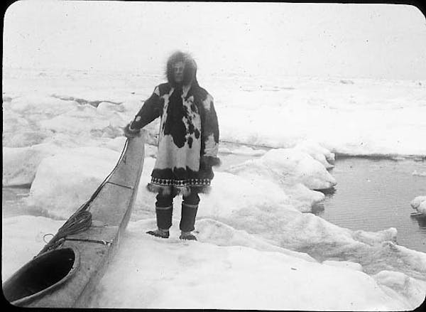 A man stands on ice with a kayak. He's wearing an animal skin coat.