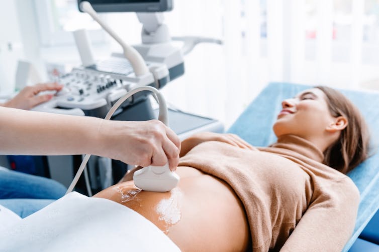 Woman having abdominal ultrasound in clinic, sonographer placing probe on belly