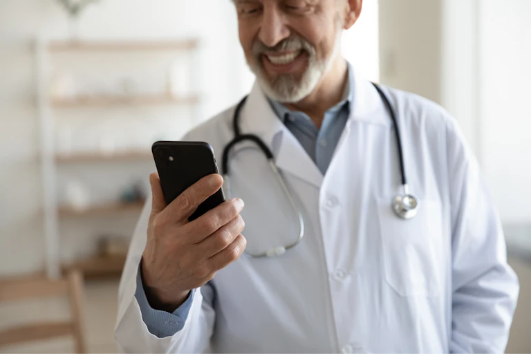 a man in a white lab coat with a stethoscope around his neck smiles at a mobile phone in his hand