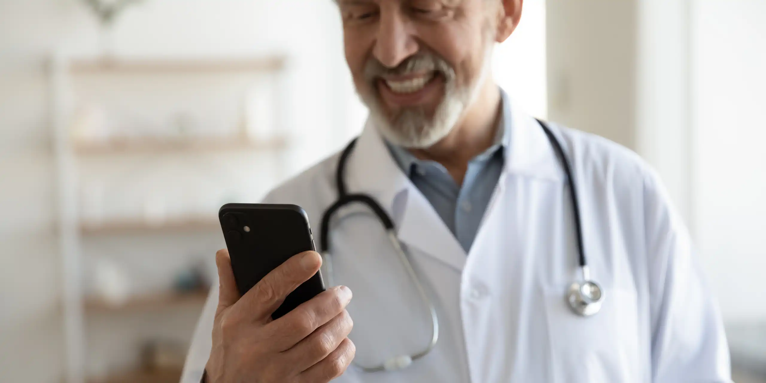 a man in a white lab coat with a stethoscope around his neck smiles at a mobile phone in his hand