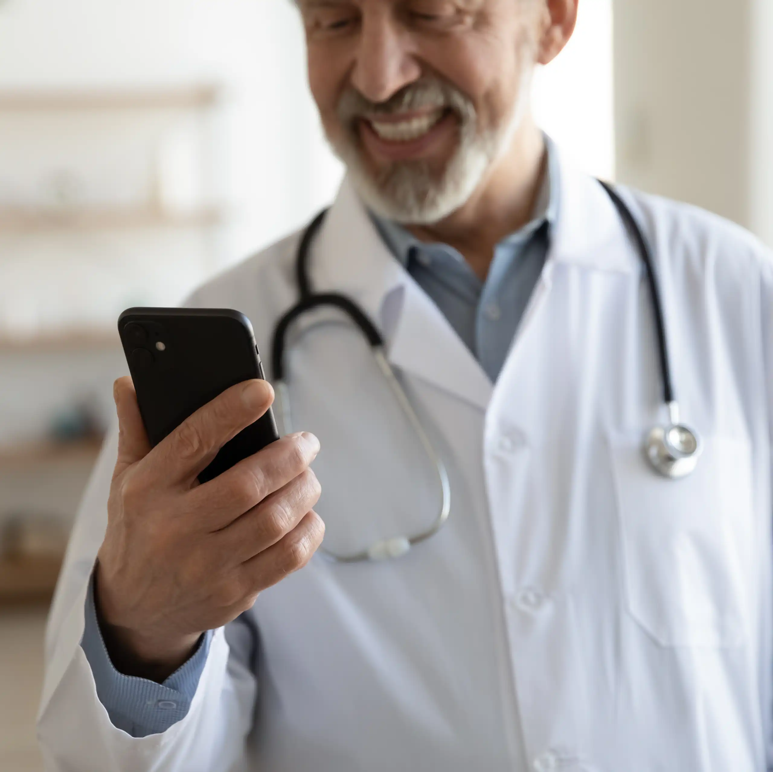 a man in a white lab coat with a stethoscope around his neck smiles at a mobile phone in his hand