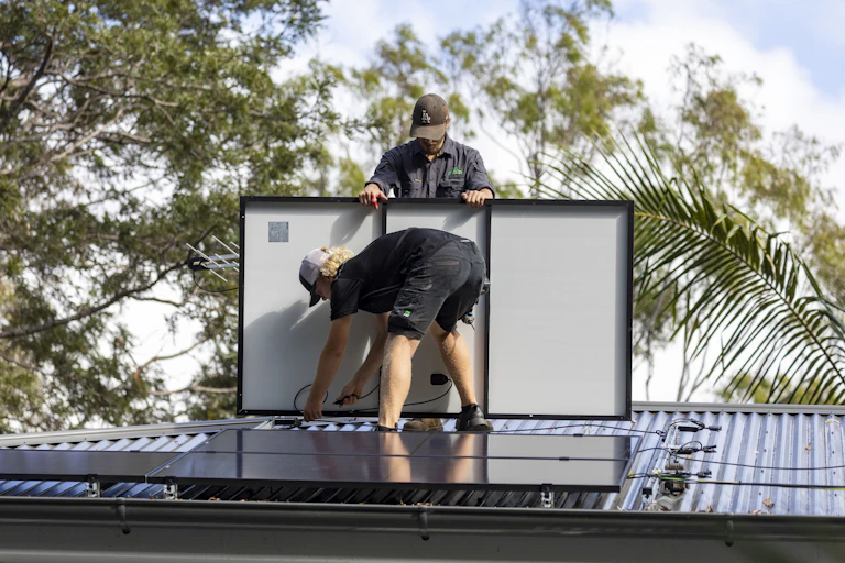 two workers on a roof installing solar panels, trees behind them