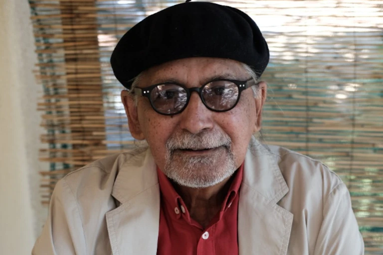 An elderly man in a beret looks directly to camera, his glasses reflecting the room he sits in.