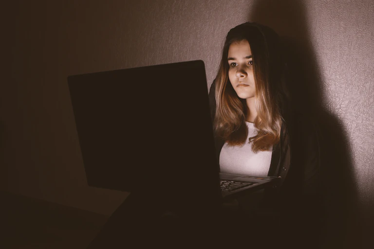 A teenage girl sits alone in a dimly lit room, her face lit up by the light of her laptop.
