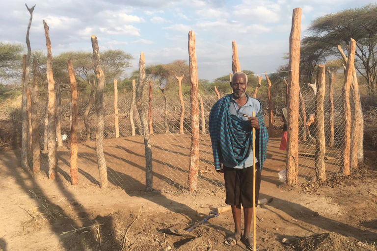 An older African man stands in front of a fenced enclosure holding a walking stick.