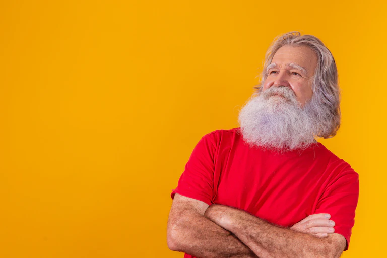 A man with a large grey beard stands with arms folded against a yellow background.