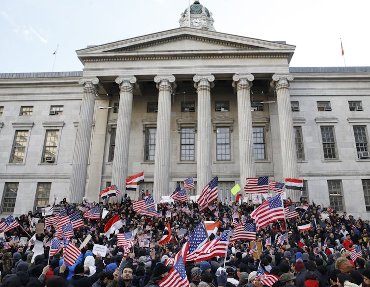 Hundreds of people waving flags gather in front a building with columns.