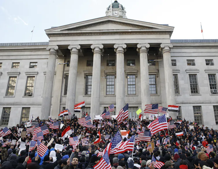 Hundreds of people waving flags gather in front a building with columns.