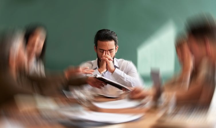 Man looking fatigued at table surrounded by blurred colleagues.