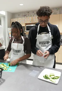 Young people wearing aprons chopping cucumbers in a kitchen.