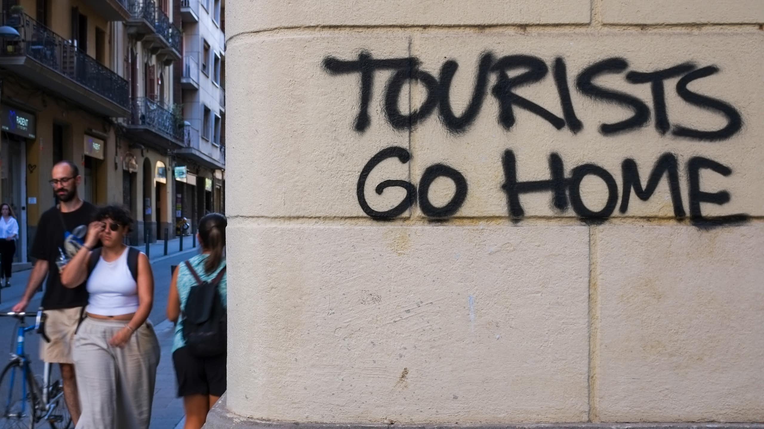 People walking down the street, passing by a graffiti on a wall that reads "Tourists go home" in Barcelona