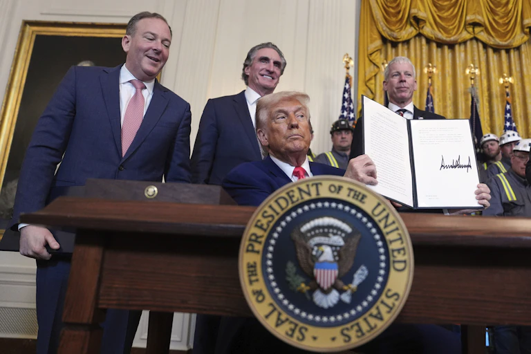 Trump sits at a desk with the seal of the president in front of it. He's holding up a folder with a signed document. Three men stand behind him smiling.