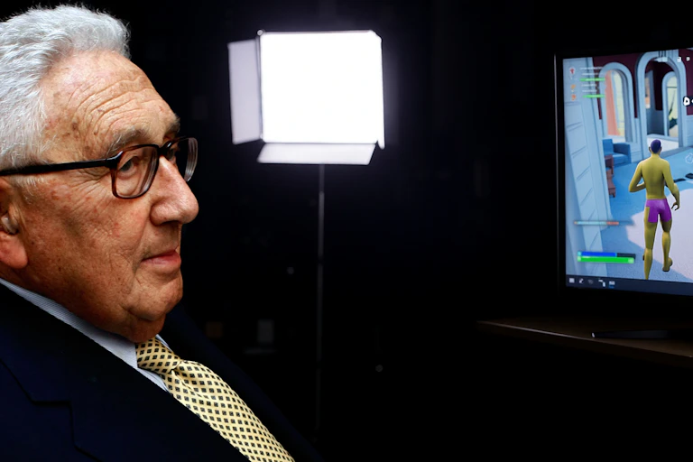 A man in glasses sits next to a screen showing a video game