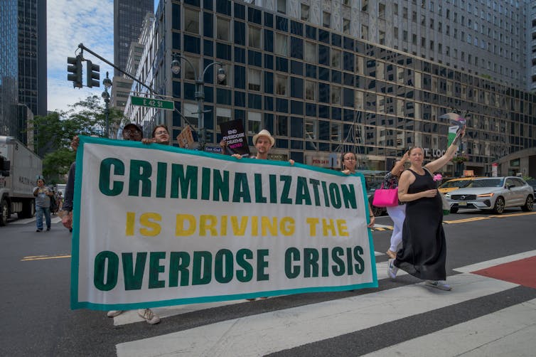 People walk down a busy street on a sunny day holding a large sign that says: