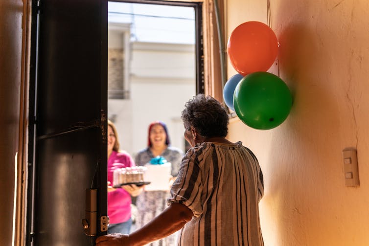 Person opening door to two people bearing cake and a present, balloons hanging on the ball