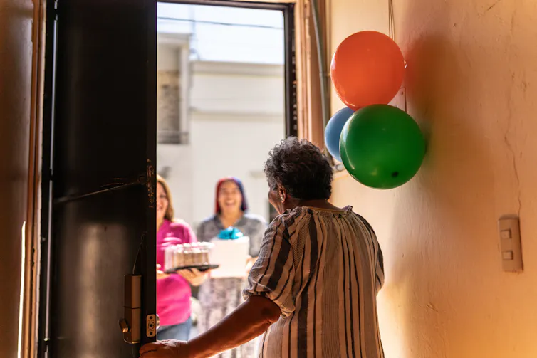 Person opening door to two people bearing cake and a present, balloons hanging on the ball
