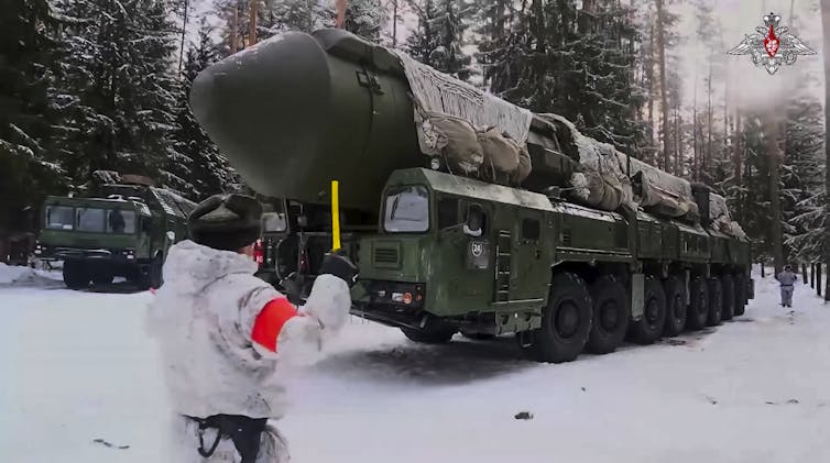 a soldier in a white uniform guides a large wheeled vehicle carrying a large missile over snow-covered ground