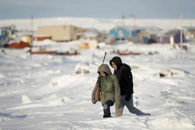 Two people in winter coats, one carrying a rifle, walk across sea ice.