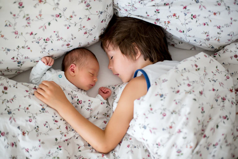 Little boy sleeping in bed next to newborn sibling.
