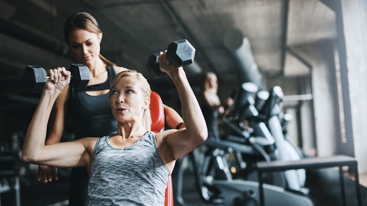 A woman lifts weights while being supervised by a trainer.