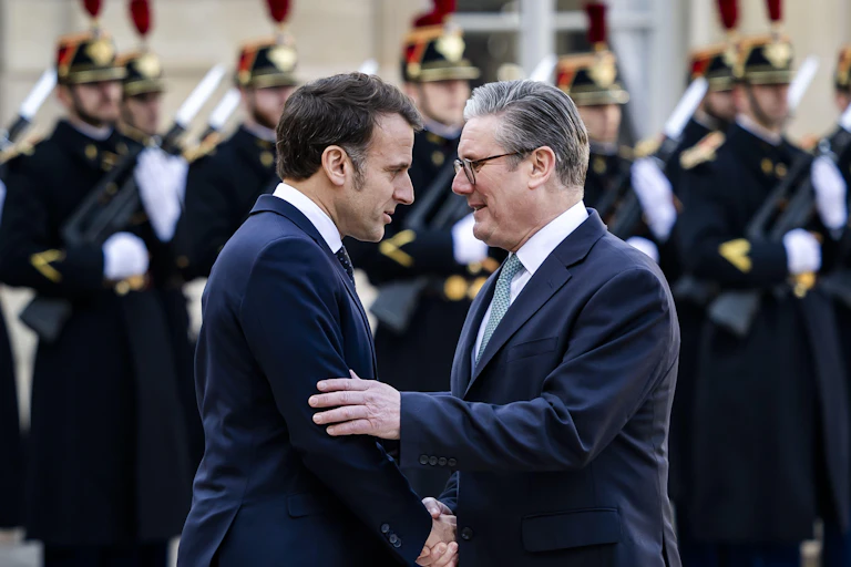 Keir Starmer and Emmanuel Macron share a close handshake with formal French military guards in the background