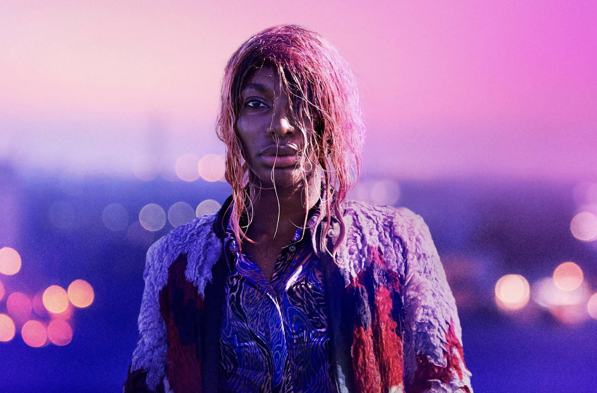 A dishevelled young black woman against a pink horizon of blurred lights looking grave.