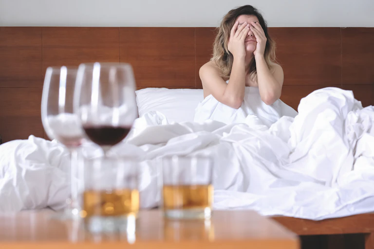 Une jeune femme assise dans un lit se tient la tête dans les mains, avec des verres à moitié vides au premier plan.