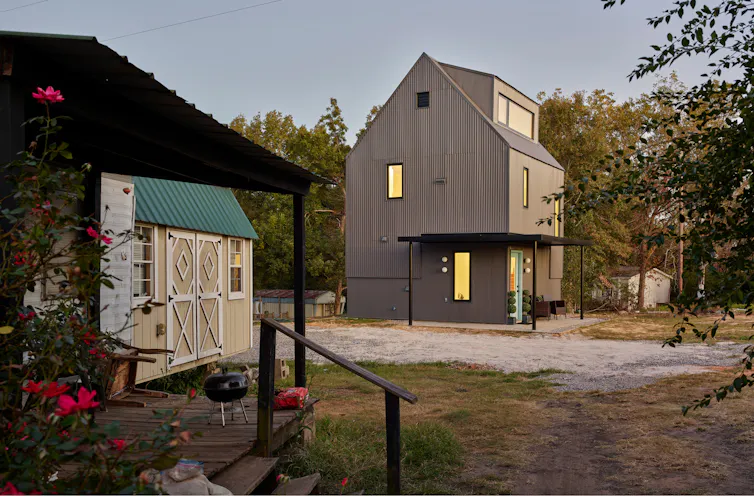 A newly built two-story home with an older small home and a utility shed in the foreground.