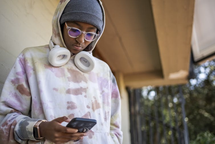 A teen in a tie-dye sweatshirt, hat and white headphones looks down at their cell phone.