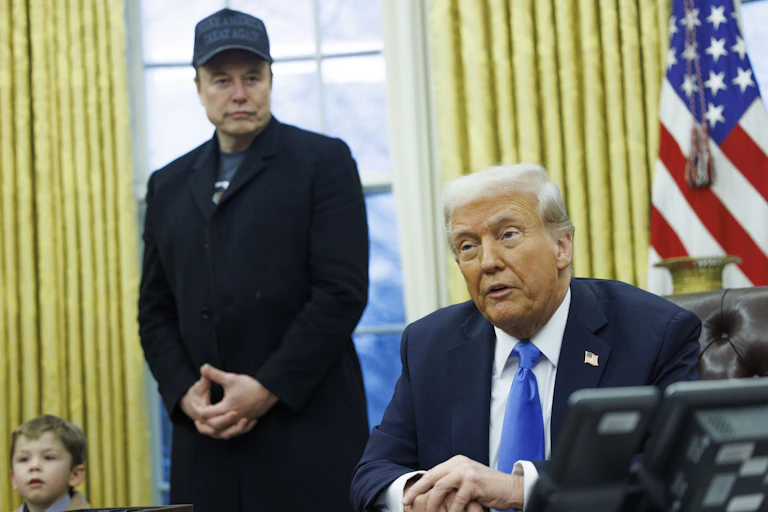 Elon Musk stands next to Donald Trump in the Oval Office. Trump is seated at a desk.