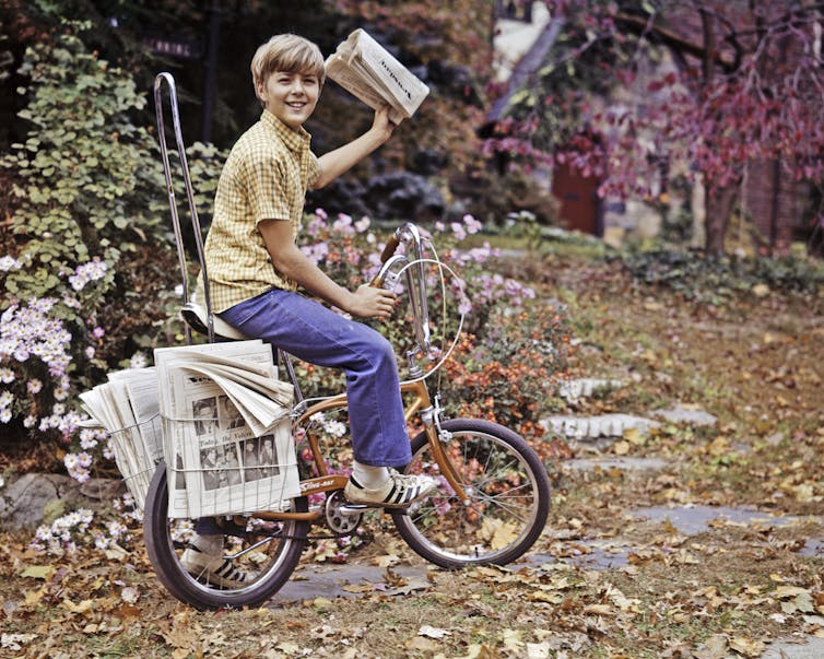 A boy with a yellow shirt and blue pants is seated on a bicycle, smiling, and has a newspaper in his hand.