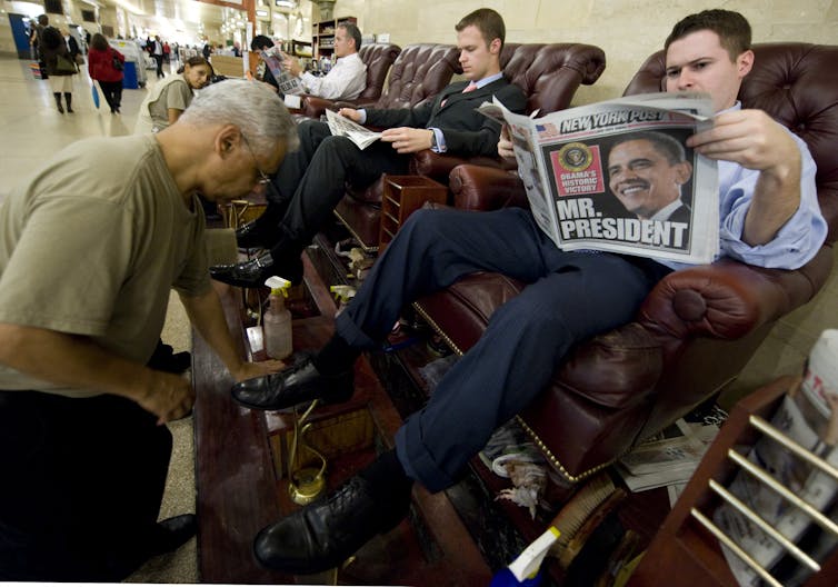 A man sits in a tall chair and holds a newspaper that shows a picture of Barack Obama and says Mr. President. A man shines the newspaper reader's shoes.