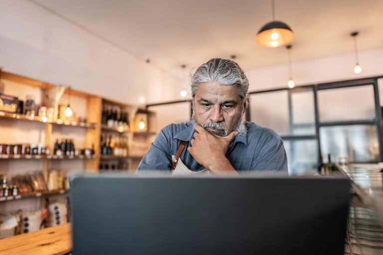 A worried-looking male business owner strokes his beard as he looks at a laptop.