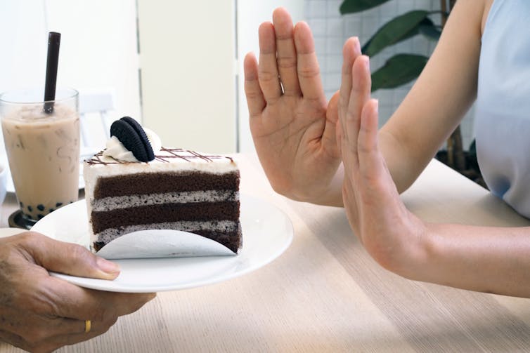 Two hands held up saying no to a proffered slice of chocolate cake.