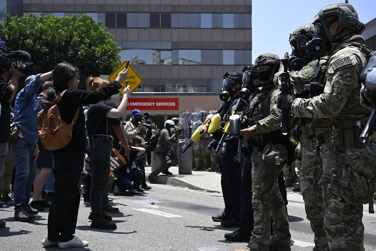 Heavily armed soldiers confront rows of protesters filming the troops with smartphones.
