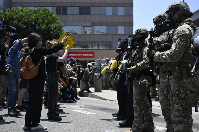 Heavily armed soldiers confront rows of protesters filming the troops with smartphones.