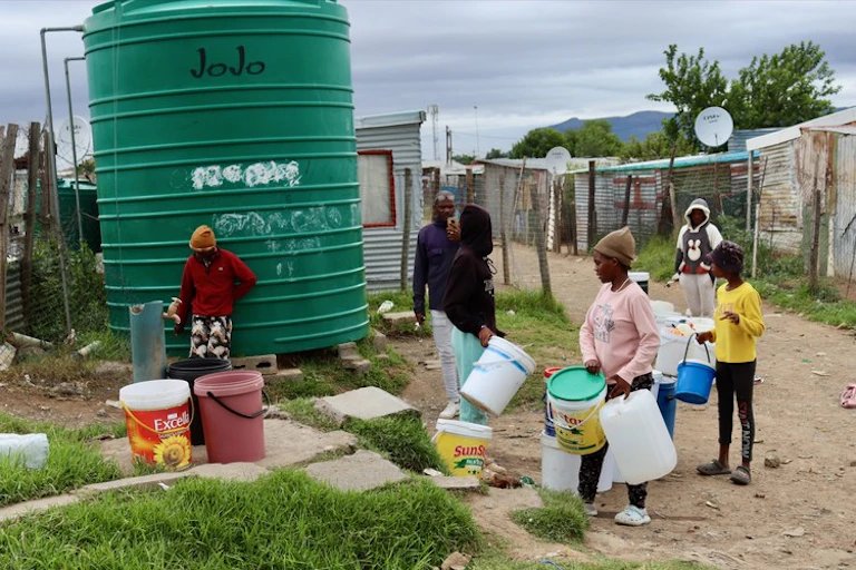 Children line up on a dusty road with buckets at a water storage tank