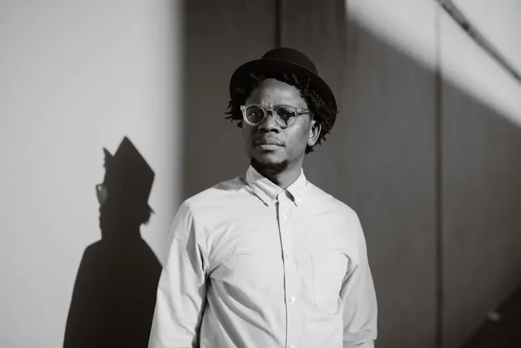 A young African man in a hat and glasses poses for a black and white portrait, his shadow cast on the wall behind him.