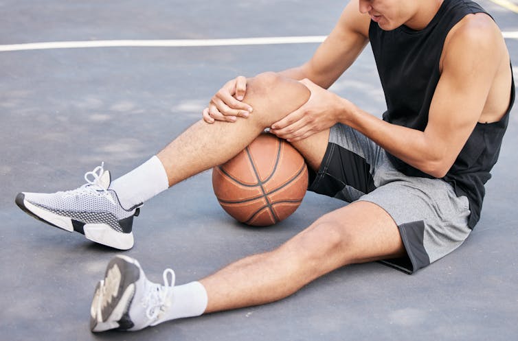 A man is sitting on the ground that is resting his injured knee on basketball.