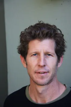 A man looks directly to camera. He has thick wavy hair and stubble on his jaw.