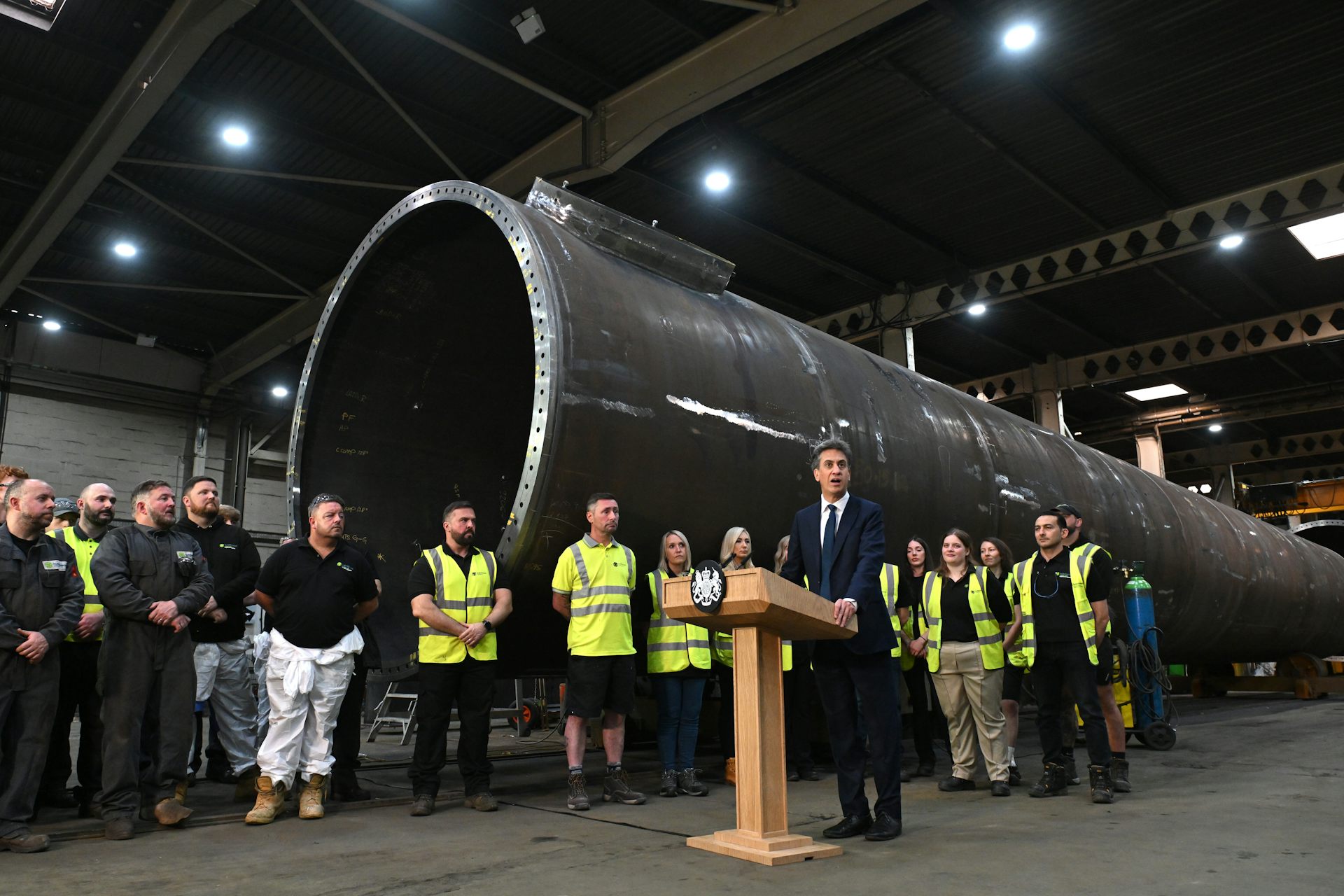 Ed Miliband at a lectern with a wind turbine component behind him.