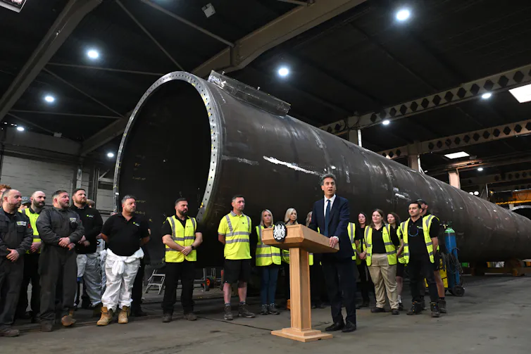 Ed Miliband at a lectern with a wind turbine component behind him.